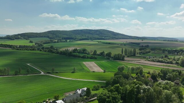 Aerial view of green fields and farmland with a forested hill and scattered houses under a blue sky in summer