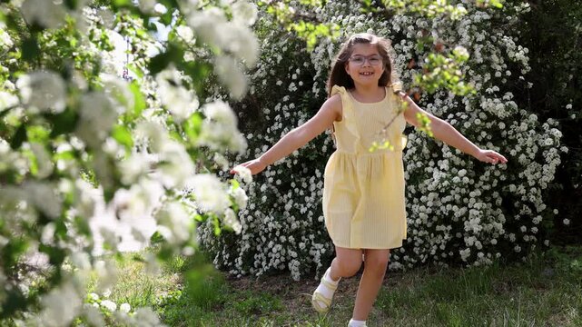 Happy little girl in yellow dress running towards camera near blossoming white spirea bushes