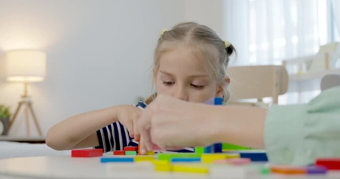 Low angle close view of preschool daughter placing colorful blocks on table while caucasian single mother assists, teaching counting, shape, balance, coordination, playful early learning at home.