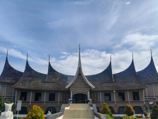Obraz premium Symmetrical view of a traditional Rumah Gadang building featuring dramatic curved roofs against a cloudy sky. Culturally authentic architecture for travel, heritage, and tourism marketing.