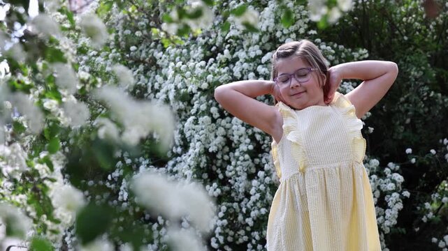 Happy little girl with glasses relaxing and holding hair behind head near blossoming white spirea bushes