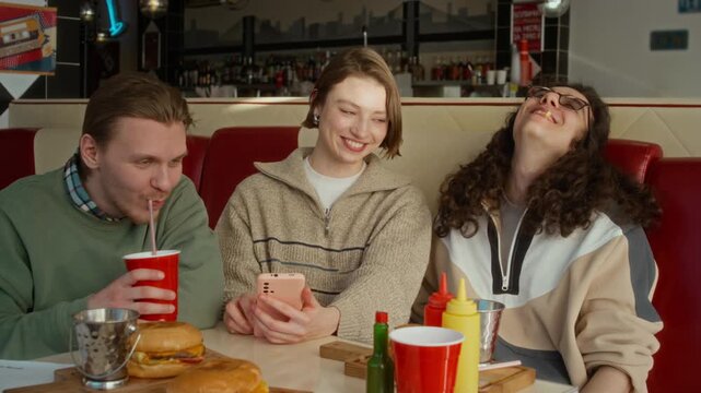Medium shot of three young college students checking out fun memes on social media using smartphone, while sitting together at cafe, laughing while enjoying hamburgers and chips