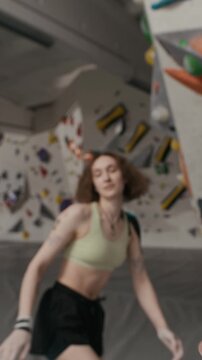 POV of young cheerful woman bouldering on indoor climbing wall, jumping down and fist bumping camera operator. Suits fitness campaigns, handheld vertical shot