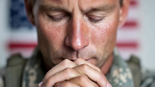 Static shot of soldier praying with hands clasped and head bowed in front of blurred american flag background inside indoor barracks close up portrait
