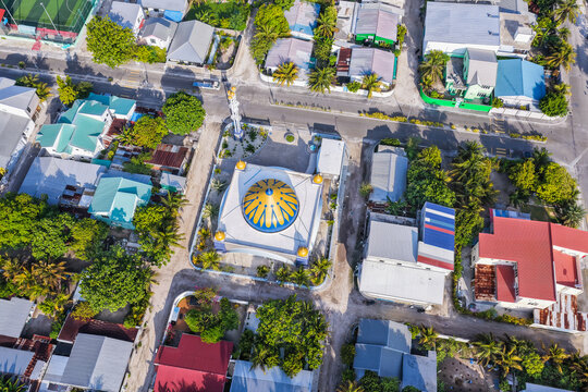 Aerial view of a mosque with a blue and yellow dome surrounded by residential houses and lush green trees in Kudahuvadhoo, Dhaalu Atoll, Maldives.