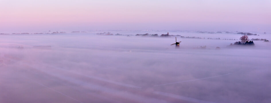 Aerial view of the historic De Vierwinden drainage mill emerging from a thick layer of morning mist over the polder landscape Opmeer, North Holland, Netherlands.