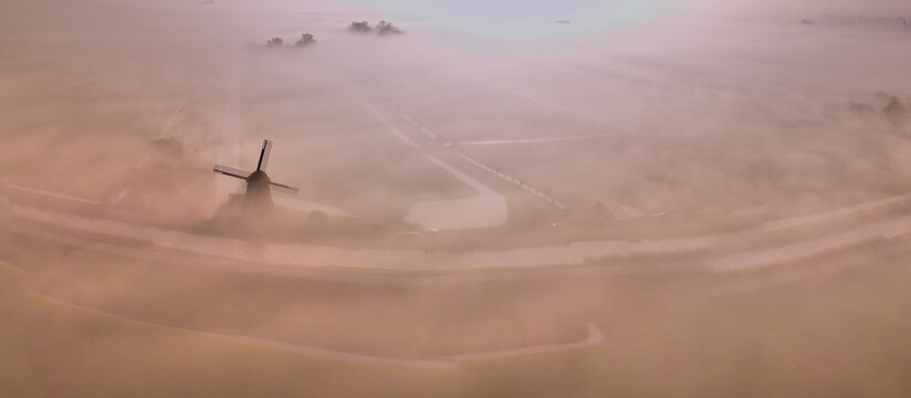 Aerial view of the historic De Vierwinden drainage mill emerging from thick morning mist over the polder landscape Opmeer, North Holland, Netherlands.