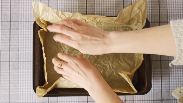 Hands of a person placing crumpled parchment paper into a baking tray, smoothing it out to fit the shape of the tray on a checkered tablecloth surface
