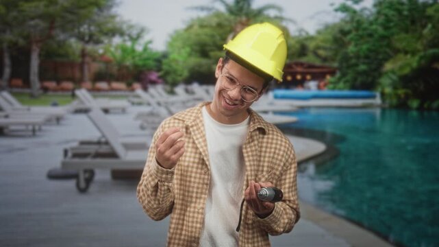 Man wearing hardhat and glasses, builder, holds cordless drill and pinches fingers while smiling near building pool deck; skill pride.