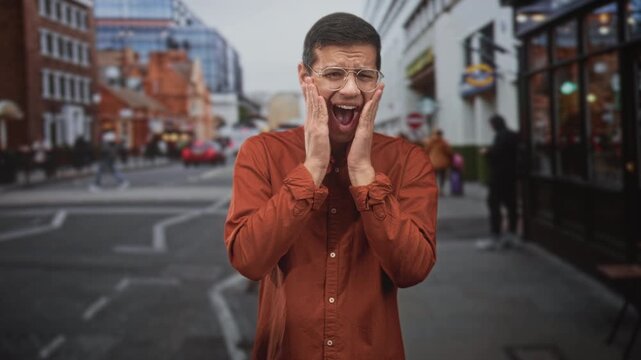 Man covering cheeks with hands, mouth open in glasses and orange shirt on busy city street with storefronts and pedestrians; surprise reaction.