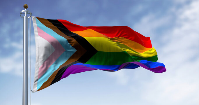 Progress Pride Flag with rainbow stripes and inclusive chevron waving from a silver flagpole against a bright blue sky with light clouds.