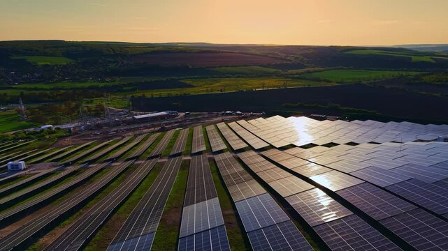 Rows of solar panels stretch across a field in a rural setting. The sun sets behind the hills. A construction site is present in the distance, showing development in the area.