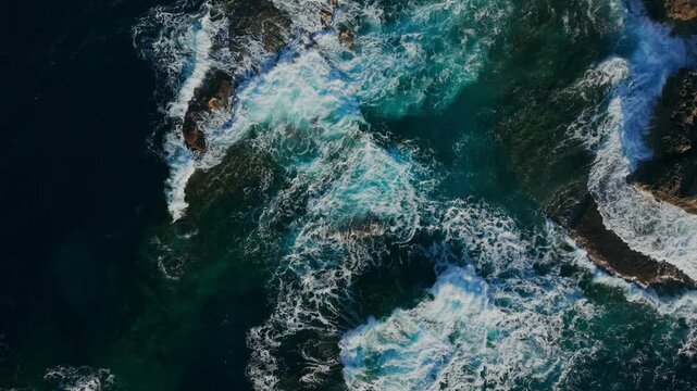Top aerial view of powerful ocean waves crashing on rocky coastline in Malta with turquoise Mediterranean water, sea foam and dramatic natural marine texture.