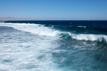 Naklejka premium Ocean waves break on a sandy beach with a clear blue sky above and distant mountains seen on the horizon