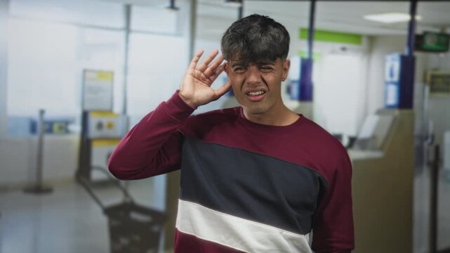 Man cupping hand to ear wincing near airport check in kiosk, facial grimace and listening gesture; confusion travel delay.