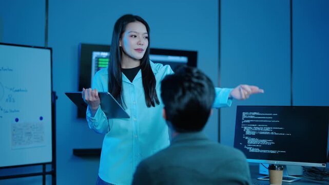 Asian female IT project manager holding a tablet discusses software development and coding with a male programmer at a computer in a modern, blue-lit tech office. Late night team collaboration.