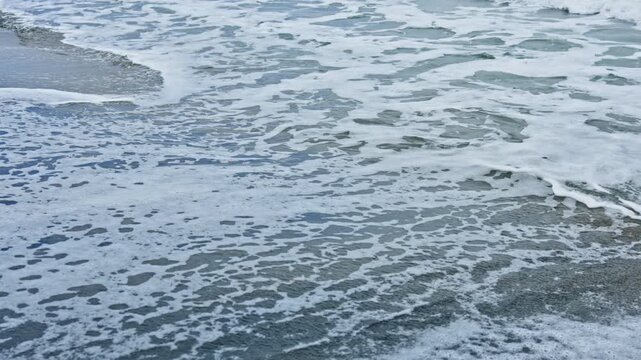 Closeup shoreline foam lapping wet sand, glittering blue reflections, gentle ebbing waves, foamy bubbles scattered across dark granular beach, ideal