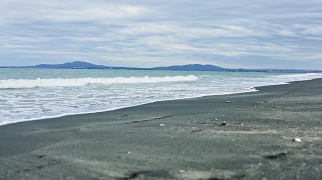 Waves roll onto the beach while the sky is covered with clouds. Distant hills are visible beyond the water, creating a natural background in this coastal setting.