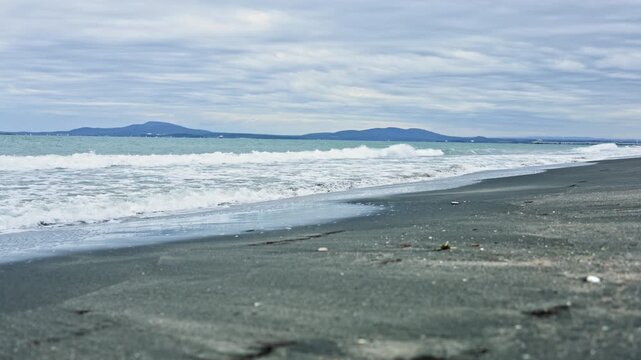 Calm black sand beach with waves under cloudy sky, distant islands on horizon, gentle surf lapping wet shore, scattered pebbles and driftwood, moody overcast