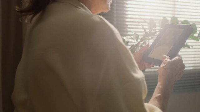 Tracking medium shot of senior Caucasian woman standing by window while holding framed vintage photo of herself and reminiscing in cozy sunlit apartment