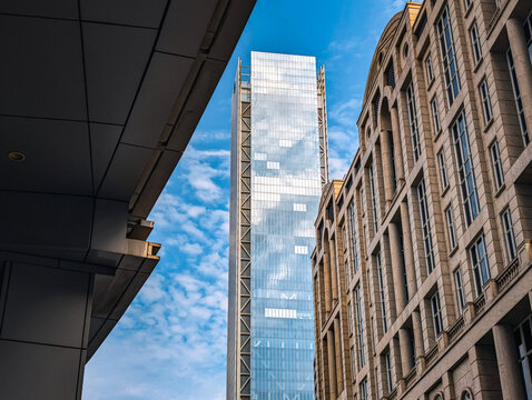 Low-angle view of a modern glass skyscraper reflecting a blue sky with scattered clouds, framed by contemporary urban architecture in a bustling metropolitan business district.
