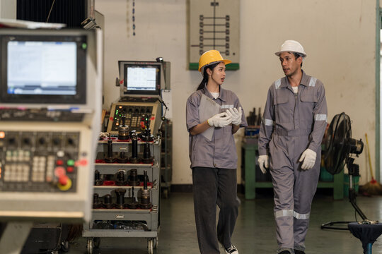 Asian woman technician and adult male operator walk past CNC station, discussing production progress, machine condition, shift coordination, and connected factory process review on workshop floor