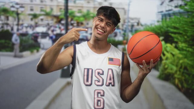 Young hispanic man points finger to cheek while holding basketball on street in front of city buildings; playful confidence.
