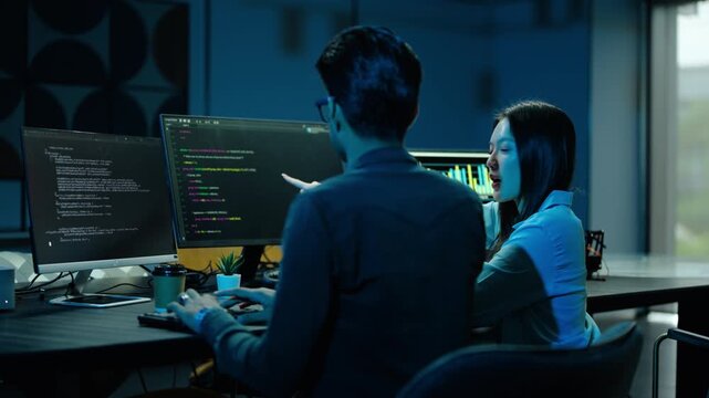 Two professional software engineers collaborating on an IT project late at night in a modern workspace, pointing at screen code and data analytics while discussing technical solutions together