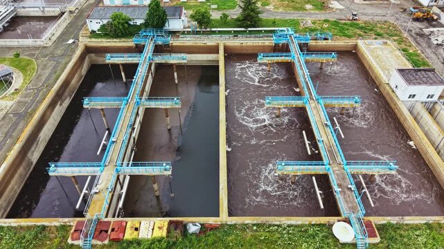 Aerial wastewater treatment plant with aeration tanks, bubbling clarifiers, concrete basins, blue mechanical aerators churning brown water, operator walkway,