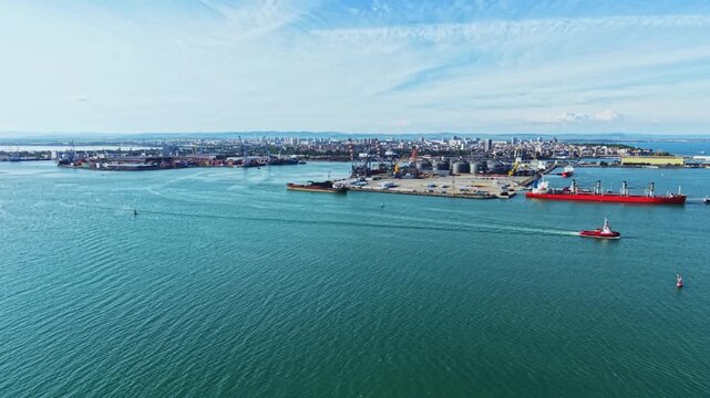 Aerial port with tugboat escort guiding tanker into busy terminal, cinematic drone footage of industrial operations, container cranes and shipyard