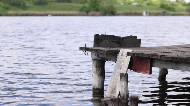 Old wooden pier on a calm lake under a cloudy sky. Traditional fishing spot, homemade wooden bridge over the water. Fishing spot on the lake shore on a sunny day. Rural landscape with green hills