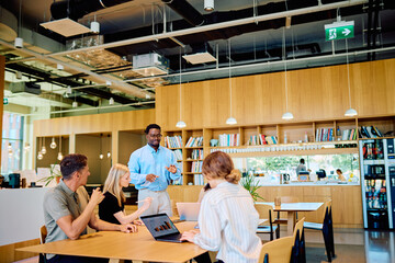 Naklejka na ściany i meble Diverse business team meeting in a modern open-plan office. A male leader stands, presenting ideas to colleagues holding laptops