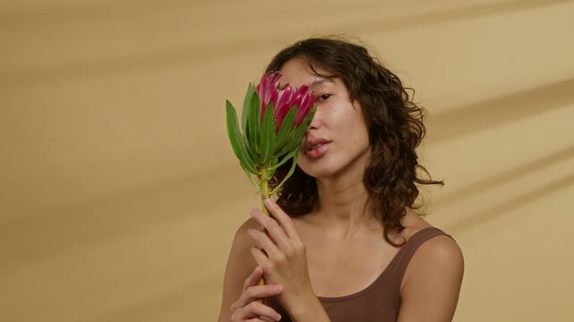 Chest-up portrait of brunette Caucasian young woman with wavy hair in brown tank top posing for camera hiding half of her face behind protea flower