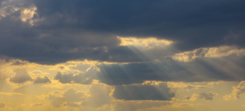Bright sunbeams effectively break through dense dark clouds during a picturesque sunset. Warm golden light illuminates the edges of cumulus clouds, creating a dramatic contrast in the evening sky.