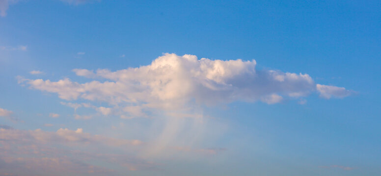 A single fluffy white cloud floats across a clear blue sky in the rays of the evening sun. Light pastel shades illuminate the bottom of the cloudlet, creating a soft and calm scenery.