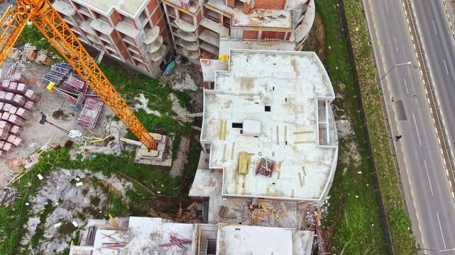Aerial top view rooftop construction beside highway, curved concrete slab and parapet form, crane boom lowering materials, material stacks and rubble