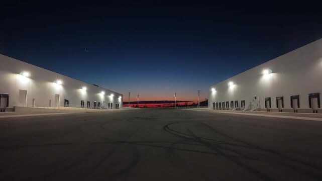The video shows an industrial area at dusk with empty loading docks and a clear sky. The lights of the building shine brightly against the darkening blue sky