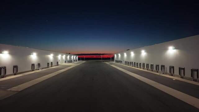 The video shows a long empty road with bright lights along the sides. The sky is darkening as the sun sets, creating a clear view of the warehouse area