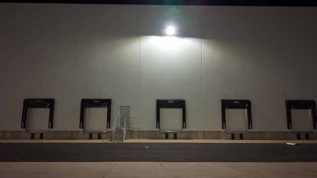 A row of loading docks at a warehouse during the night. Bright lights shine on the wall above. Empty spaces under the lights are visible