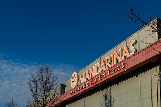 Vilnius, Lithuania - March 3 2026: Exterior of Mandarinas Shopping Center glowing in warm evening sunlight, bold signage on modern facade, clear deep blue sky, bare winter trees, calm weather.