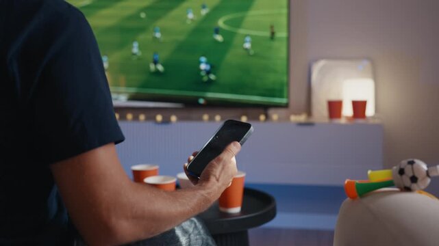 Man placing sports bet on smartphone while watching soccer match on tv