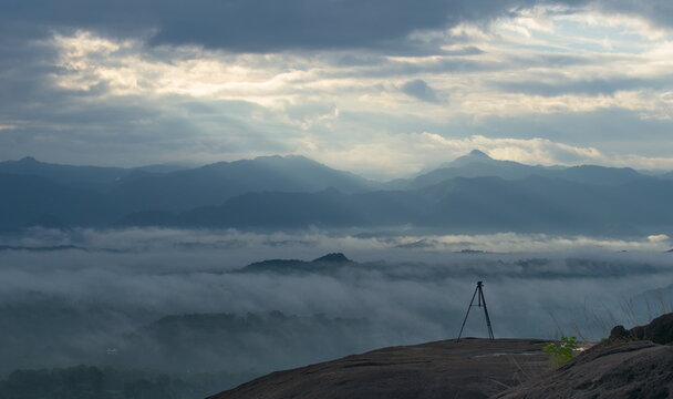 Serene valley fog and rolling hills under a cloudy sky with crepuscular rays, featuring a camera tripod set up on a rock edge for landscape photography.