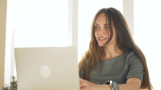 Young woman uses laptop computer. Smiling woman sits by bright sunny window. Working online brings positive emotion. Freelance worker enjoys typing. Happy student finishes online working task.