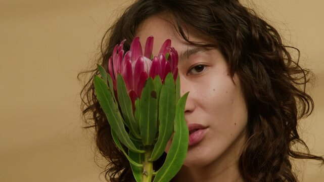 Close-up portrait of young woman with brown eyes looking at camera holding pink protea flower in front of her face