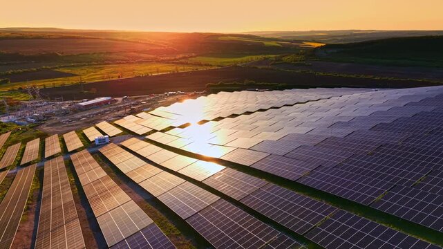 The scene shows numerous solar panels arranged neatly in rows across a large field. The sun sets in the background, casting warm colors over the landscape.