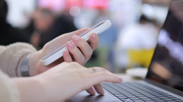 Hands hold phone while typing on laptop. Person multitasks, focused and engaged. Background blurred with soft lighting. Ideal for work-from-home, remote collaboration, or digital multitasking themes