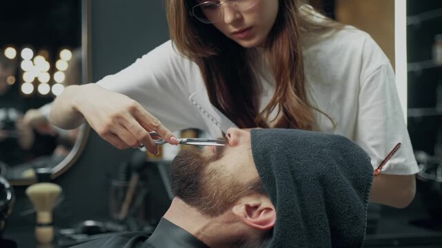 Professional barber trimming beard of male client with scissors and comb during grooming procedure in modern barbershop salon