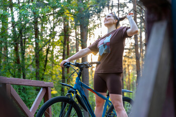 Naklejka premium Woman Stretching On Wooden Bridge With Bicycle At Sunset, Warm Golden Light Through Pines, Silhouette And Confident Pose, Quiet Training Moment