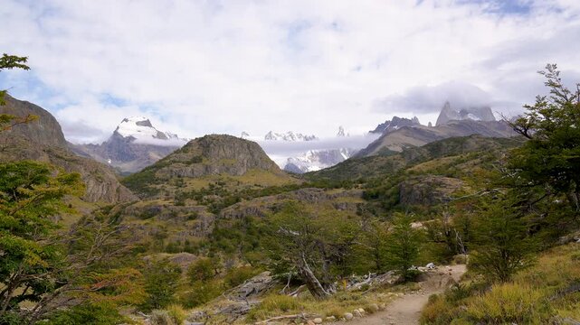 Scenic hiking trail in El Chalten leading  towards the majestic snow-capped peaks of Fitz Roy in Patagonia, surrounded by green trees and rocky hills under a cloudy sky