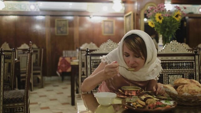 Slow motion shot of female traveler tasting traditional soup at authentic restaurant in Cairo, Egypt. Woman in headscarf enjoying local Middle Eastern cuisine in warm oriental atmosphere captured in c